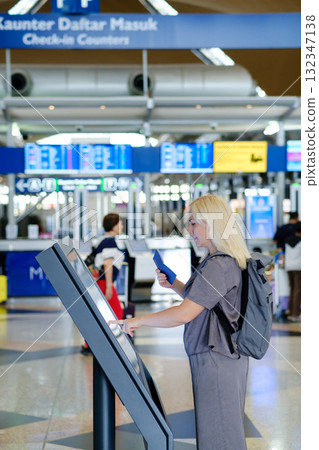 Woman checks in at an airport self-service kiosk while traveling, with busy terminal in the background during afternoon hours 132347138