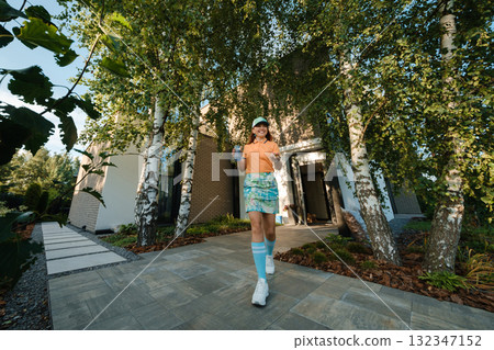 Young person walks towards modern house surrounded by greenery, showcasing a bright exterior and well-maintained garden in the afternoon light 132347152