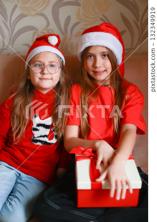 Girls in New Year's looks with a gift are sitting at home on the sofa. Girls in New Year's looks with a gift are sitting at home on the sofa. 132349919