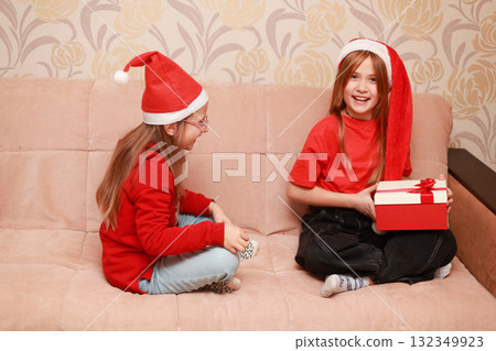 Girls in New Year's looks with a gift are sitting at home on the sofa. Girls in New Year's looks with a gift are sitting at home on the sofa. 132349923