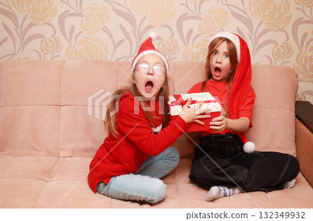 Girls in New Year's looks with a gift are sitting at home on the sofa. Girls in New Year's looks with a gift are sitting at home on the sofa. 132349932