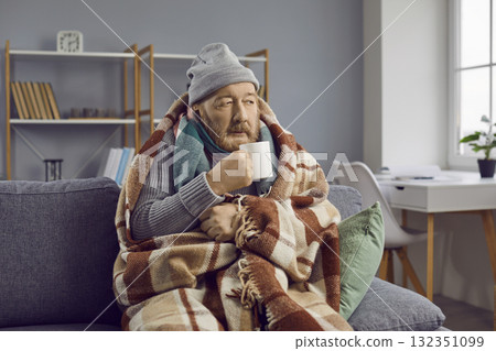 Senior man warms up and treats himself with hot drink while sitting on sofa at home. Senior man warms up and treats himself with hot drink while sitting on sofa at home. 132351099