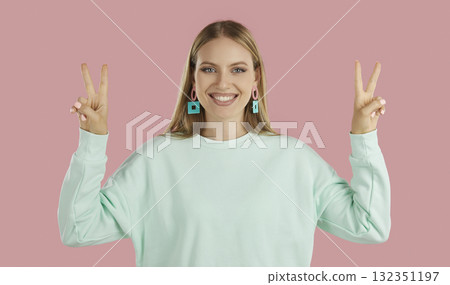 Blondy happy girl in mint sweatshier showing victory signs by two hands on pink background. Blondy happy girl in mint sweatshier showing victory signs by two hands on pink background. 132351197