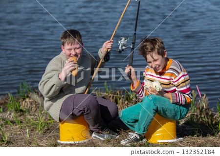 The boys are friends on a fishing trip by the river eating a bun and laughing. Children are friends on a summer fishing trip. The boys are friends on a fishing trip by the river eating a bun and laughing. Children are friends on a summer fishing trip. 132352184