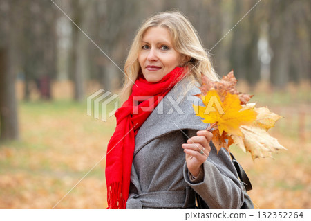 Beautiful adult blonde woman with autumn leaves. She is wearing a gray coat and a red scarf. 132352264