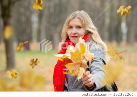 An adult woman in a red scarf and a gray coat shows a bouquet of yellow maple leaves. 132352265