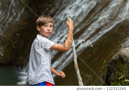 A tourist boy in a white shirt with a stick against the background of a blurred waterfall with a mountain. 132352268