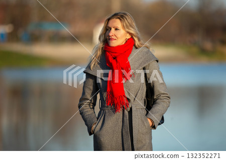 Beautiful adult blonde woman in a gray coat against the background of a lake. Beautiful adult blonde woman in a gray coat against the background of a lake. 132352271