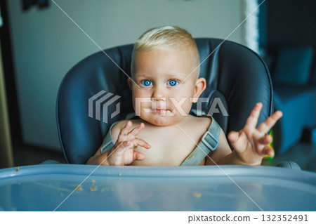A little baby boy is sitting in a high chair. An emotional child sits in a chair and eats. Feeding a child. 132352491