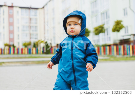 A little boy in overalls runs on the street. A child in a raincoat walks in the yard. Children's overalls are mudproof 132352503