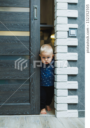 Blond toddler boy at home peeks into the room through a crack in the door. Boy 1 year old opens the door to the room. Children at home. 132352505
