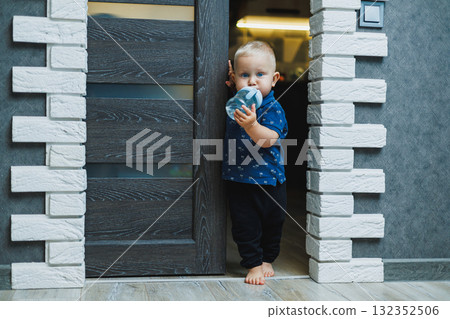 Blond toddler boy at home peeks into the room through a crack in the door. Boy 1 year old opens the door to the room. Children at home. 132352506
