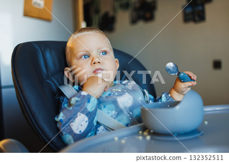 Cute one-year-old boy sits in a high chair and eats independently. Silicone utensils for feeding children. 132352511