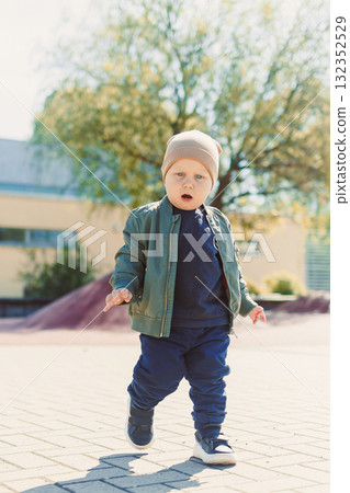 A 2 year old boy in a hat and jacket runs and plays outside. Toddler on a spring walk in the park. Child outdoors on a playground. A 2 year old boy in a hat and jacket runs and plays outside. Toddler on a spring walk in the park. Child outdoors on a playground. 132352529