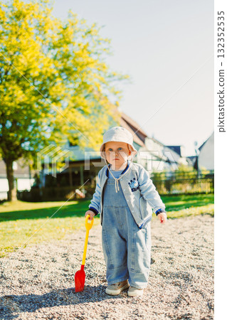 Toddler playing with a shovel in the sandbox on the playground. Boy playing in nature. 2 year old boy enjoying green grass in the park on a sunny summer day. 132352535