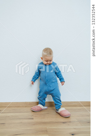 Little toddler boy in a blue romper tries on big house slippers. Child plays at home in the living room. Children's home clothes. 132352544