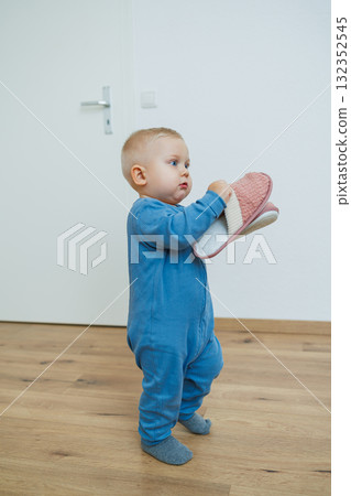 An adorably happy toddler dressed in a cozy, colorful outfit is gleefully holding a pair of slippers while surrounded by a bright and cheerful interior space filled with joyful decor and furniture An adorably happy toddler dressed in a cozy, colorful outfit is gleefully holding a pair of slippers while surrounded by a bright and cheerful interior space filled with joyful decor and furniture 132352545