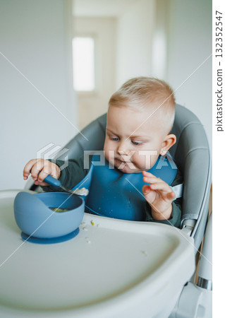 Blue-eyed toddler boy sitting on a chair with a silicone apron eating independently. Toddler boy learning to eat independently. Safe utensils for feeding children. 132352547