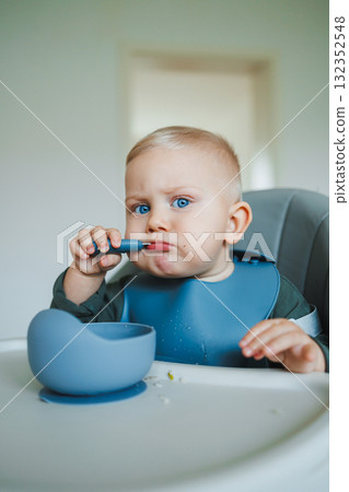 An Adorable Cute Baby is Joyfully Enjoying Mealtime, Eagerly Using a Blue Spoon and Bowl, Sitting Comfortably in a High Chair, Creating a Mess in a Happy, Playful Atmosphere of Family Life An Adorable Cute Baby is Joyfully Enjoying Mealtime, Eagerly Using a Blue Spoon and Bowl, Sitting Comfortably in a High Chair, Creating a Mess in a Happy, Playful Atmosphere of Family Life 132352548