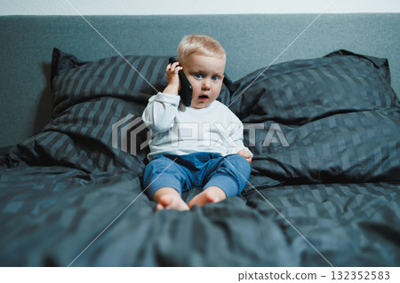 Handsome toddler boy in a white t-shirt and jeans sits on the bed and watches cartoons on the phone. 2 year old boy plays with a mobile gadget. Modern digitalization of toddlers. Handsome toddler boy in a white t-shirt and jeans sits on the bed and watches cartoons on the phone. 2 year old boy plays with a mobile gadget. Modern digitalization of toddlers. 132352583