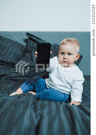 Handsome toddler boy in a white t-shirt and jeans sits on the bed and watches cartoons on the phone. 2 year old boy plays with a mobile gadget. Modern digitalization of toddlers. Handsome toddler boy in a white t-shirt and jeans sits on the bed and watches cartoons on the phone. 2 year old boy plays with a mobile gadget. Modern digitalization of toddlers. 132352585