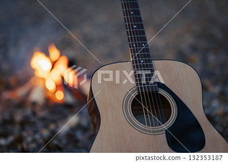 Close-up shot of acoustic guitar near bonfire outdoors. New guitar standing near burning campfire in the evening, spending time outdoors on nature Close-up shot of acoustic guitar near bonfire outdoors. New guitar standing near burning campfire in the evening, spending time outdoors on nature 132353187