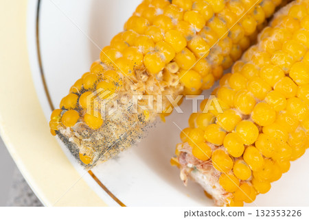 Yellow corn kernels on plate with signs of decay and deterioration. Couple of yellow spoiled corn cobs on the plate, showcasing a mix of untouched kernels and areas affected by mold 132353226