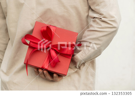 Man hiding red gift box tied with ribbon behind their back, wearing beige shirt for surprise girlfriend. Birthday present suggests surprise or special occasion. Valentine's Day. Close up 132353305
