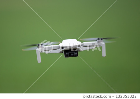 Front view of a flying drone with a camera. Flying small consumer quadcopter on background of green field Front view of a flying drone with a camera. Flying small consumer quadcopter on background of green field 132353322