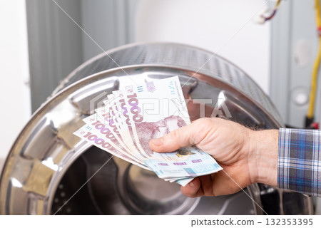 Hand holding hryvnia money near disassembled washing machine in repair setting. Hand holding Ukrainian hryvnia banknotes in front of a disassembled washing machine, indicating a repair payment Hand holding hryvnia money near disassembled washing machine in repair setting. Hand holding Ukrainian hryvnia banknotes in front of a disassembled washing machine, indicating a repair payment 132353395