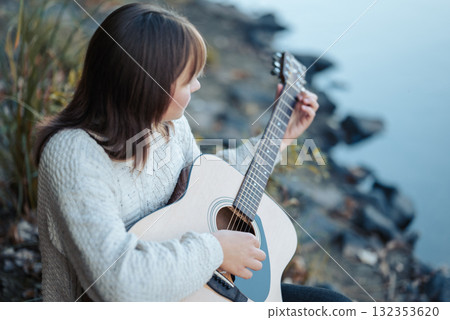 Outdoor portrait of young girl sitting on the bank of the river and playing the guitar. Warm autumn evening on nature, enjoying music and calmness Outdoor portrait of young girl sitting on the bank of the river and playing the guitar. Warm autumn evening on nature, enjoying music and calmness 132353620