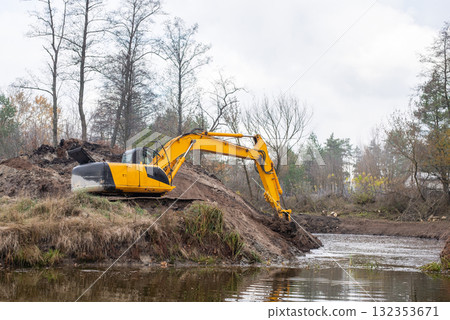 Big industrial excavator digging pond in the countryside. Yellow excavator shoveling dirt from the bottom of an artificial pond 132353671