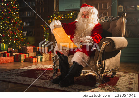 Merry Christmas. Happy Santa Claus reading wish list paper roll in festive living room with fireplace and Christmas tree. Santa with xmas wishlist letter on Christmas eve. Winter Holiday Celebrations 132354041