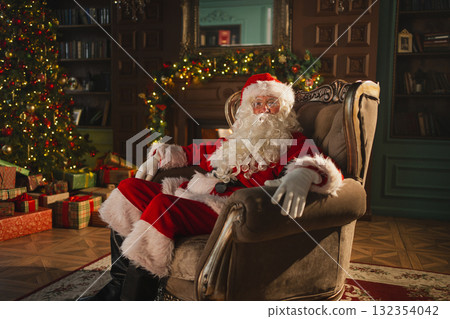 Merry Christmas. Happy Santa Claus sitting in chair in festive living room with fireplace decorations Christmas tree. Santa in cozy home on Christmas eve. Winter New Year Holiday Celebrations 132354042