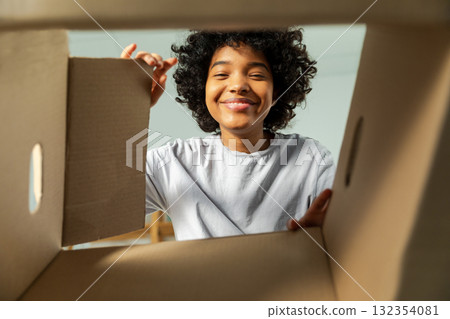 Inside of cardbox bottom view. African girl unpacking delivery looking in box. Woman opening carton box. Female getting parcel looking at delivered goods items. Satisfied client positive feedback 132354081