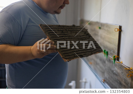 A person applies adhesive to the back of a ceramic tile before installing it on an interior wall. A close-up of the tile installation process during home renovation and improvement. A person applies adhesive to the back of a ceramic tile before installing it on an interior wall. A close-up of the tile installation process during home renovation and improvement. 132354803