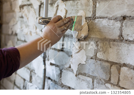 A worker manually applies putty to an old brick wall resurfacing and repairing surfaces with a putty knife and follows a laser line for alignment during a home renovation. Leveling a wall with putty. A worker manually applies putty to an old brick wall resurfacing and repairing surfaces with a putty knife and follows a laser line for alignment during a home renovation. Leveling a wall with putty. 132354805