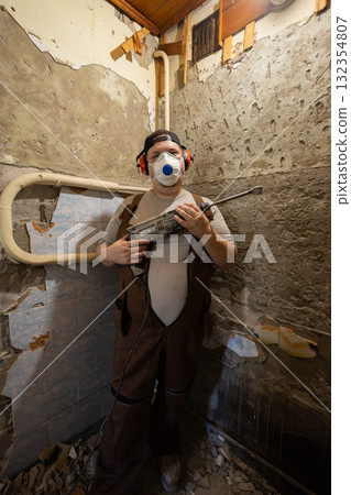 Man wearing personal protective equipment performing renovation work, standing in a room with walls stripped of old plaster and cement, holding a hammer drill. Man wearing personal protective equipment performing renovation work, standing in a room with walls stripped of old plaster and cement, holding a hammer drill. 132354807