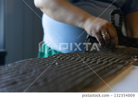 A worker applies adhesive to a tile with a notched trowel. The adhesive is evenly distributed across the rough surface of the tile. A close-up shows the texture and skill of the hands. 132354809