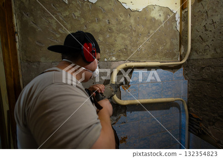 A worker prepares a wall for repair, wearing a protective mask and ear protection, and removes old plaster with a hammer drill. A man also uses a hammer drill to remove tiles from the walls. 132354823