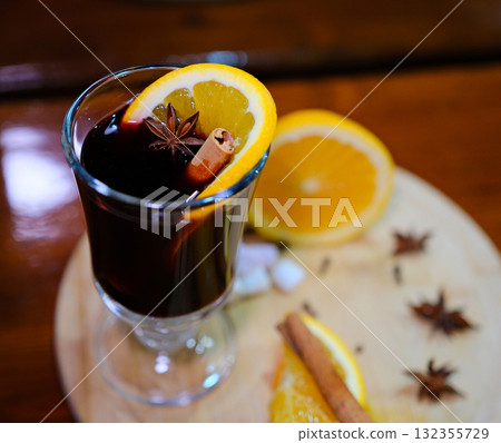 An overhead view of a glass of mulled wine, garnished with a clove-studded orange slice. The scene is set against a red background, with a wooden cutting board holding two more orange slices and loose 132355729