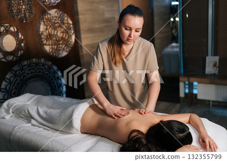 Professional massage therapist performing deep massaging technique on female client's back in serene cozy spa environment, promoting relaxation and healing. Concept of rest with spa treatment. 132355799