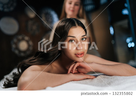 Attractive woman client getting relaxing back massage from professional massage therapist while looking at camera lying resting on table, experiencing therapeutic wellness at luxurious spa setting. Attractive woman client getting relaxing back massage from professional massage therapist while looking at camera lying resting on table, experiencing therapeutic wellness at luxurious spa setting. 132355802
