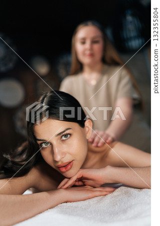 Vertical portrait of attarctive woman lying looking at camera in wellness center during massage therapy. Portrait of beautiful female client getting relaxing arm and shoulder massage in spa salon. 132355804