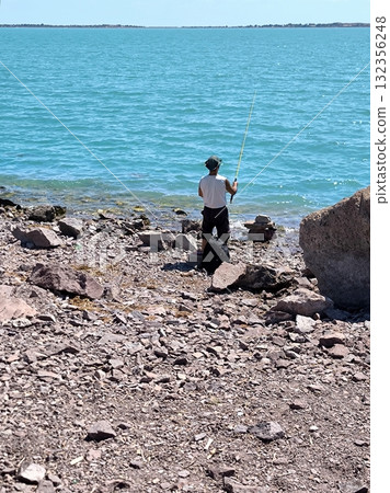 A fisherman fishes from a rocky shore against the backdrop of a turquoise lake. A fisherman fishes from a rocky shore against the backdrop of a turquoise lake. 132356248