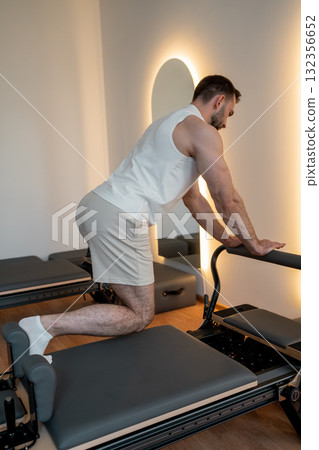 Man training pilates on the reformer bed. Reformer pilates studio machine for fitness workouts in gym. Fit, healthy and strong authentical body. Fitness concept 132356652