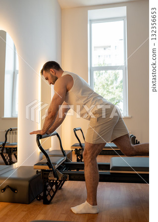 Man training pilates on the reformer bed. Reformer pilates studio machine for fitness workouts in gym. Fit, healthy and strong authentical body. Fitness concept Man training pilates on the reformer bed. Reformer pilates studio machine for fitness workouts in gym. Fit, healthy and strong authentical body. Fitness concept 132356653