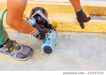 Carpenter in gloves operates pneumatic nail gun to secure wooden boards on floor of busy construction site. 132357154