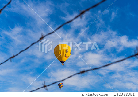 Hot air balloon with a painted smile floats in a clear blue sky, viewed through barbed wire, creating a playful yet thought-provoking travel and freedom concept Hot air balloon with a painted smile floats in a clear blue sky, viewed through barbed wire, creating a playful yet thought-provoking travel and freedom concept 132358391