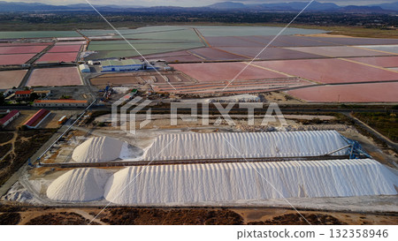 Aerial drone view of colorful salt evaporation ponds and white salt piles near Santa Pola, Spain, with industrial structures and mountains in the distance. 132358946